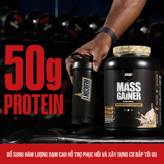 Hand holding a protein shaker and mass gainer supplement with visible text on a blurred gym background.
